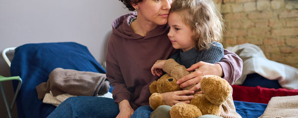 Mother embracing her young girls who is holding a teddy bear while they sit on a shelter bed.